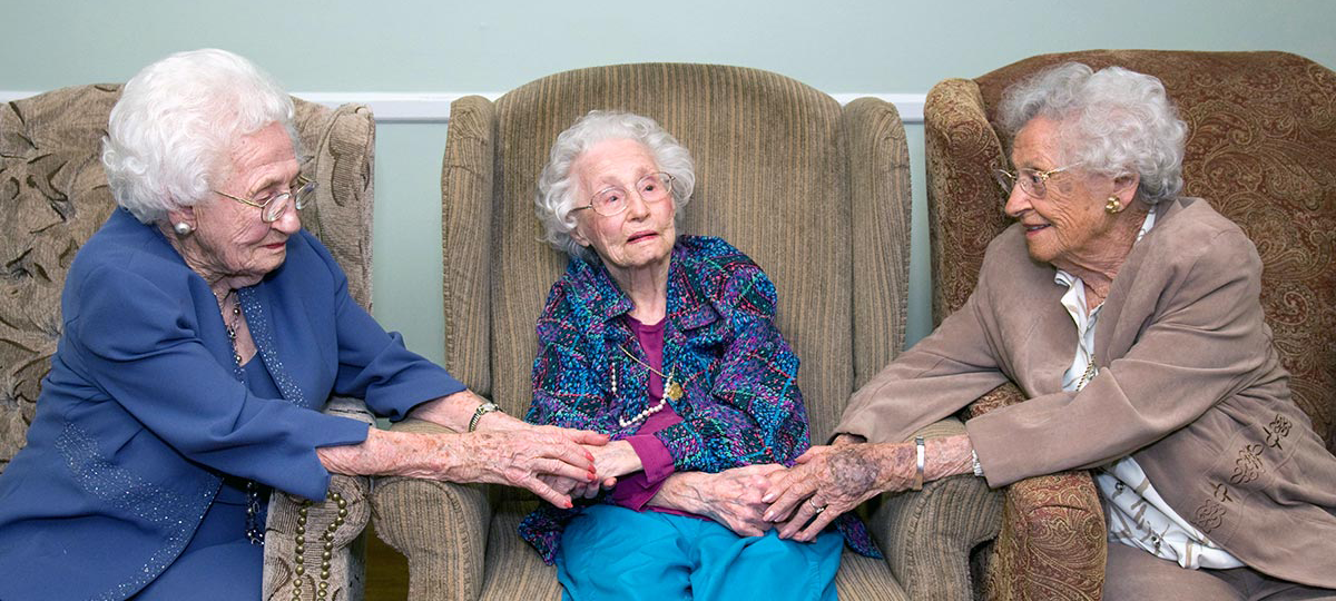 Three elderly women seated in armchairs hold hands