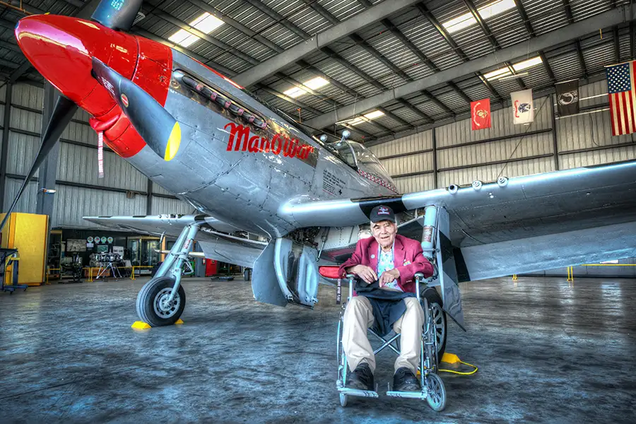 An older man in a wheelchair sits in front of a vintage airplane in a hangar
