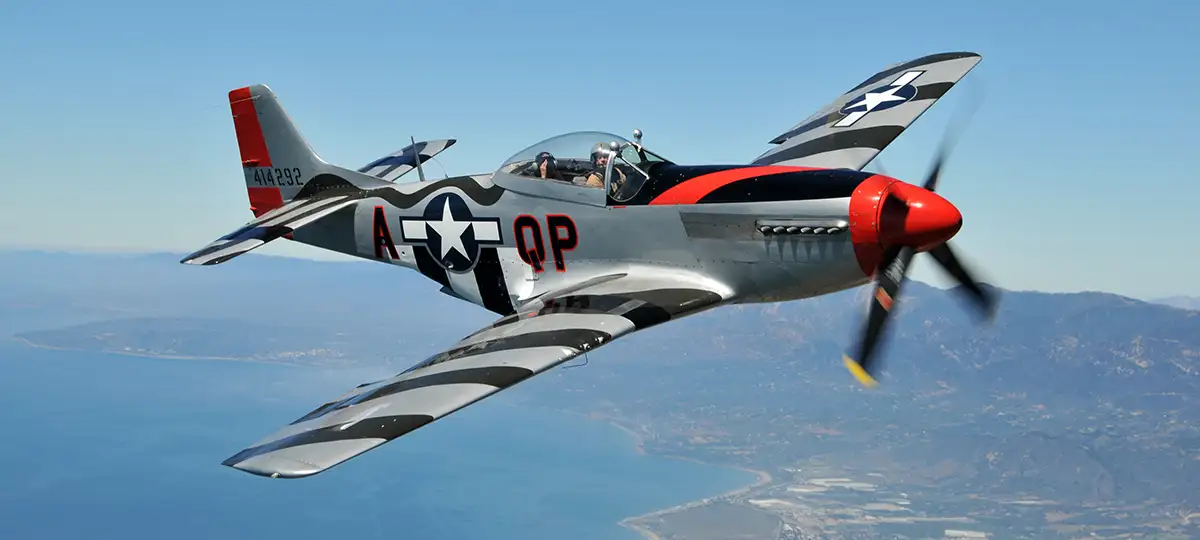 A silver, red, and black P-51 Mustang airplane flies above ocean and mountains