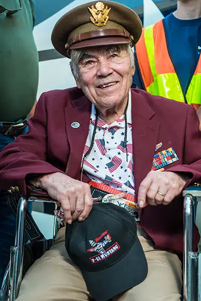 A WWII veteran wears a Commander's hat while holding a baseball cap embroidered with "P-51 Mustang"