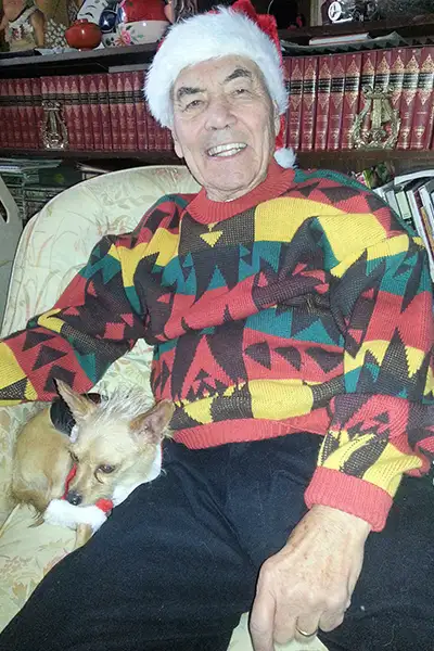An older gentleman wearing a Santa hat sits in an armchair with a tiny dog next to him