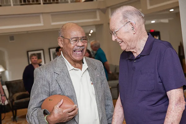 Two older men, one holding a football, laugh in conversation.