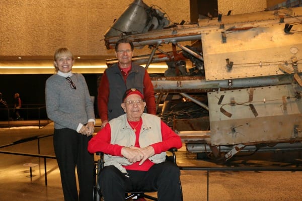A man and a woman stand behind a man in a wheelchair with wreckage from September 11, 2001, in the background