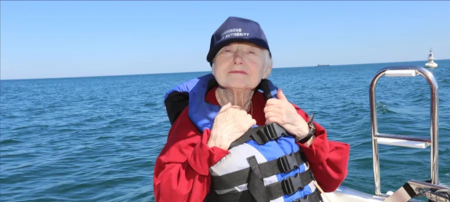 An older woman wearing a baseball cap and life jacket sits on a sailboat on the water