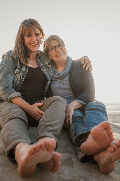 Colleen and her daughter embrace while sitting on big rock on beach with bare feet