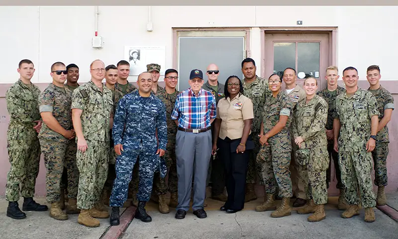 A group of men and women in Army fatigues stand with an older man wearing a veteran ballcap