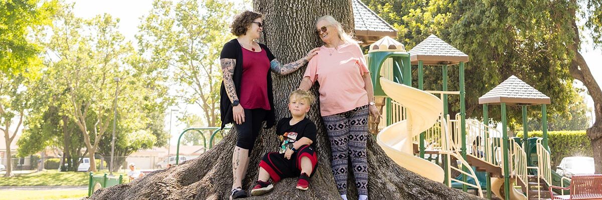 Christie, Vickie, and Brayden smile and look at each other in front of big tree in park