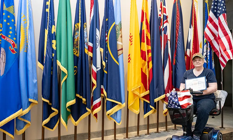 An older woman holds a certificate of honor in front of a row of flags.