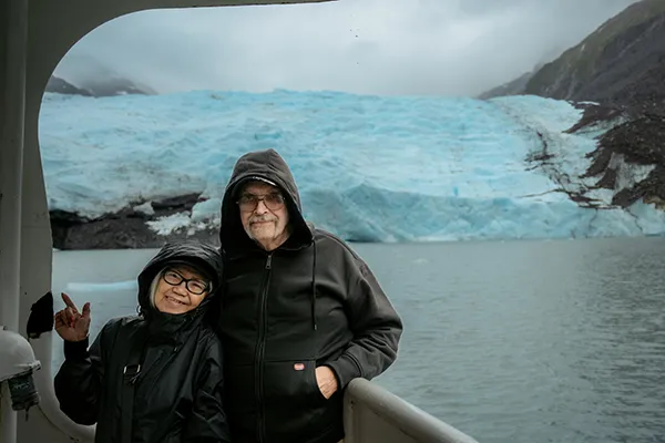 A couple stands in front of a glacier on an Alaskan boat tour.