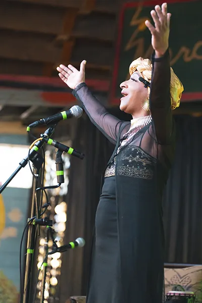 A Black woman sings into a microphone with arms raised.