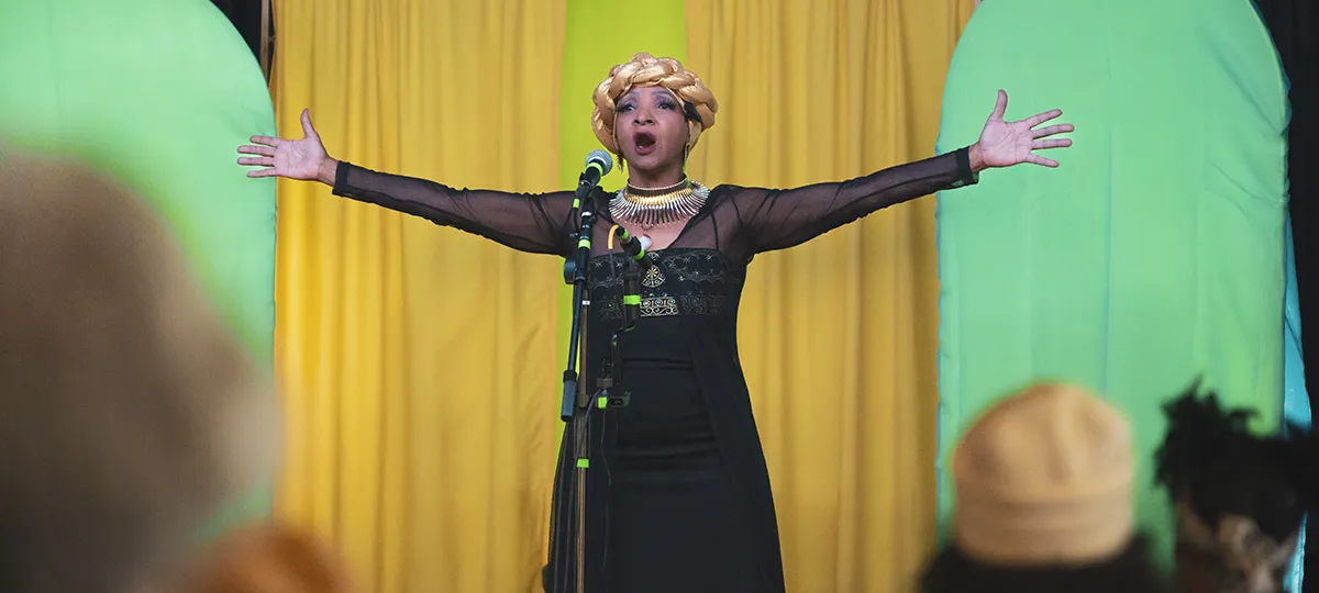 A Black woman with a head covering sings on stage with arms outstretched.