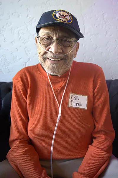 An older man smiles with an oxygen tube and Navy veteran hat.