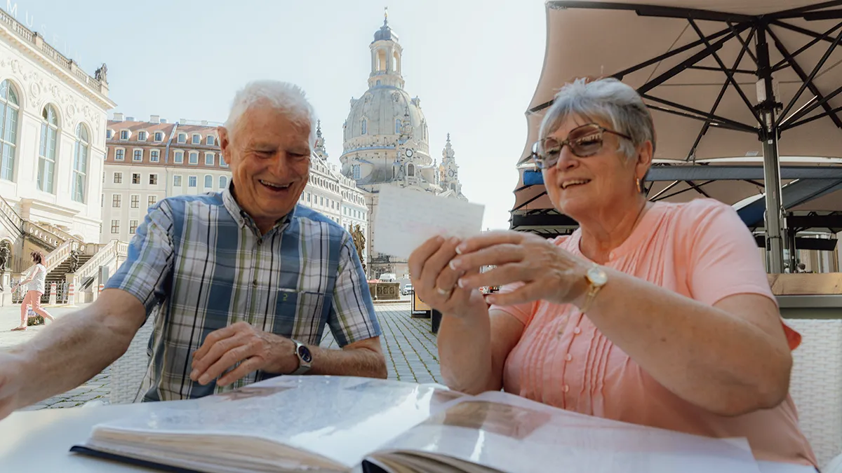 An older man and woman look at a scrapbook outdoors in a German city.