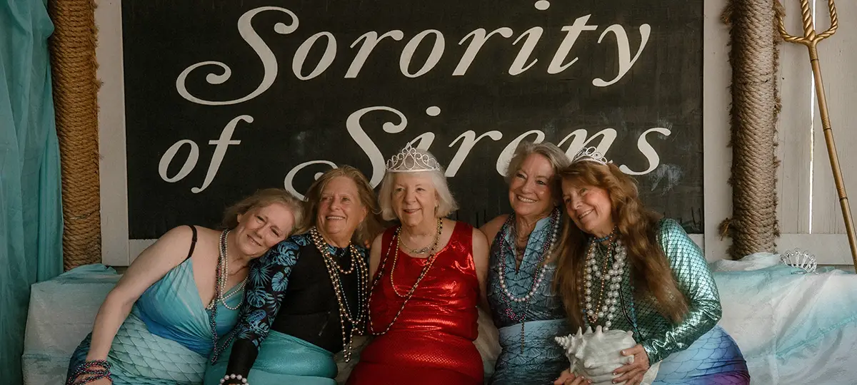 Five older women wearing beads dress as mermaids.