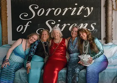 Five older women wearing beads dress as mermaids.
