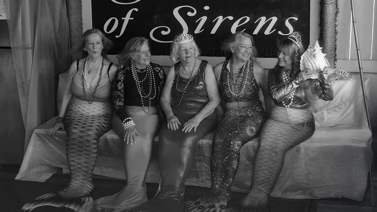 Five women wearing mermaid costumes sit on a sofa together.