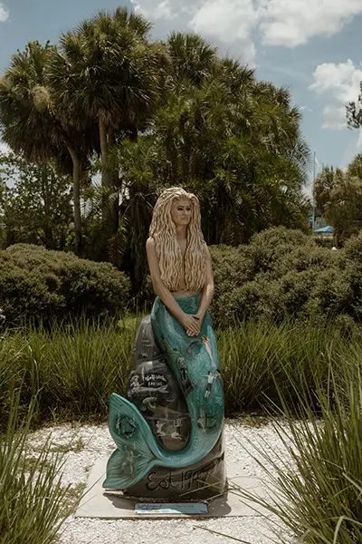 A statue of a mermaid surrounded by trees, bushes, and native grasses.