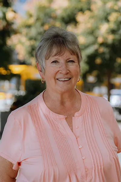 An older woman in a pink blouse smiles.