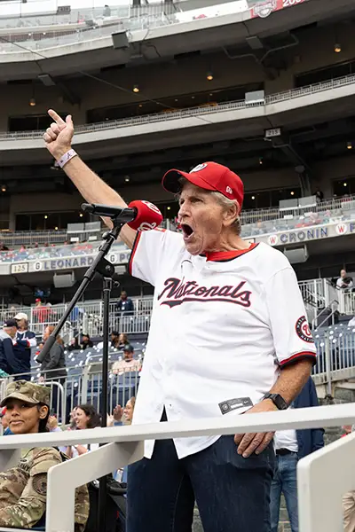 A man wearing a Nationals jersey and baseball cap shouts into a microphone in a stadium.