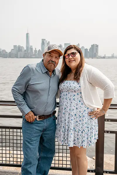 An older man stands along the water with a younger woman with a New York City in the background.