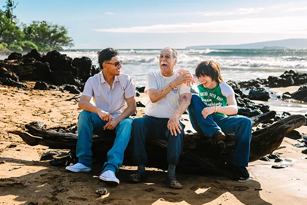 An older man and two younger men laugh while sitting on the beach.