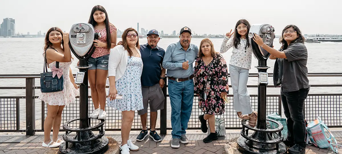 Three generations of Hispanic family members stand on a deck near water with a city in the background.