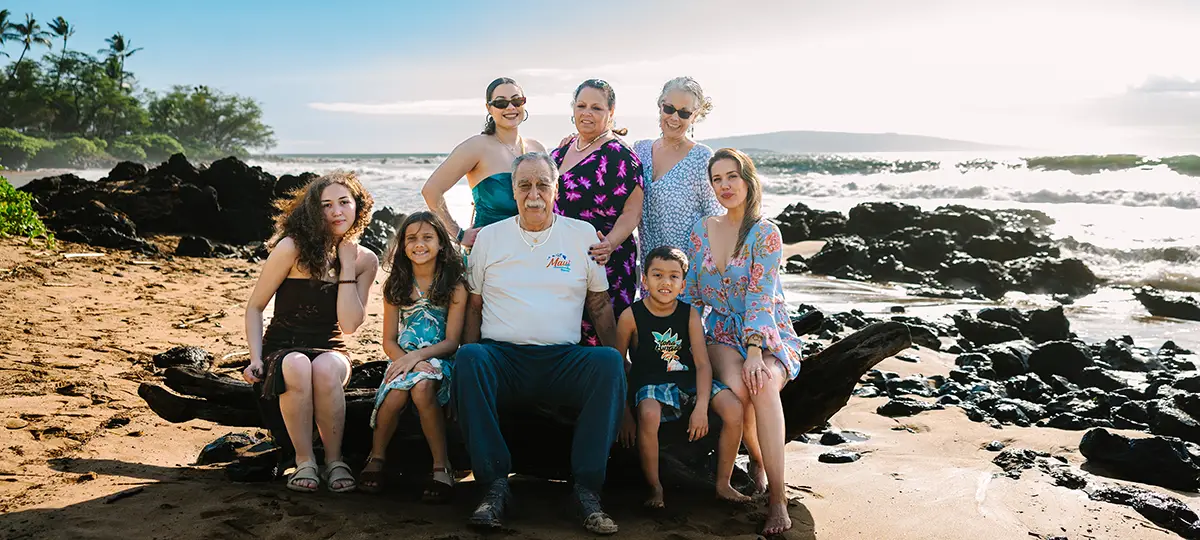 Three generations sit together on a rocky shore along the ocean.