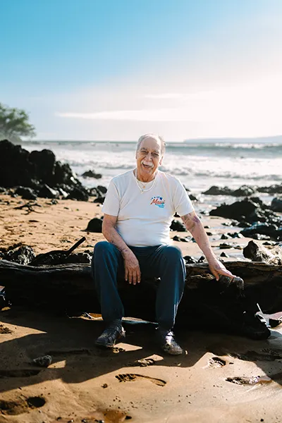 An older Mexican man sits along a rocky ocean shoreline.