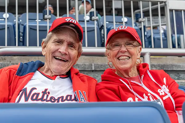 A man and woman in baseball gear smile from stadium seats.