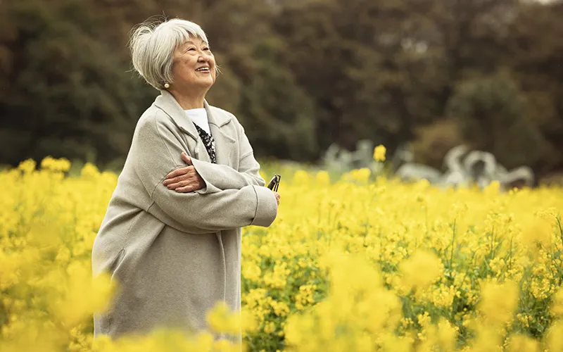 An older Japanese American woman stands in a field of yellow flowers