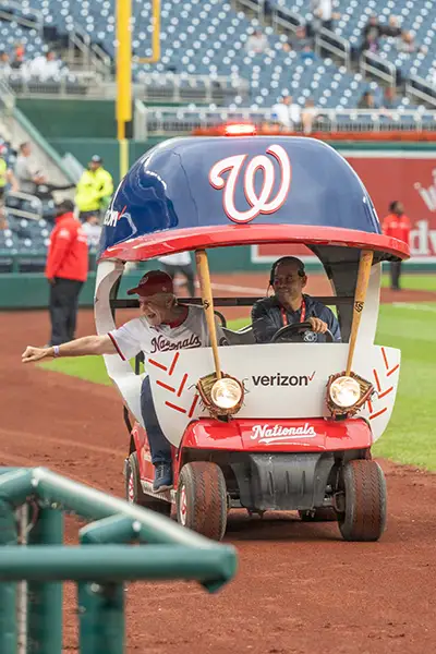Two men ride in a Washington Nationals bullpen cart on a baseball field.