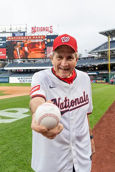 An older man in a baseball jersey holds a baseball on the Washington Nationals field.