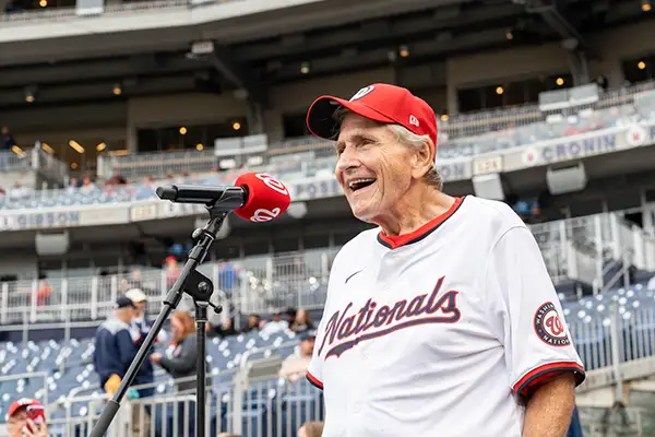 An older man in baseball gear stands at a microphone.