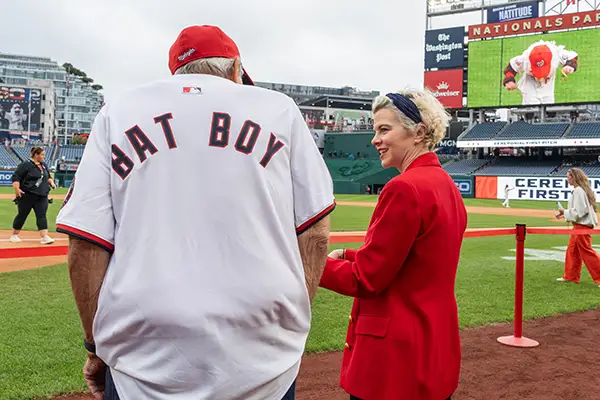 An older man in a bat boy jersey stands next to a young woman on the edge of a baseball field.