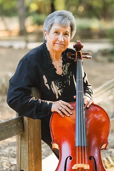 A Hispanic woman leans on a fence with her hands on a cello.