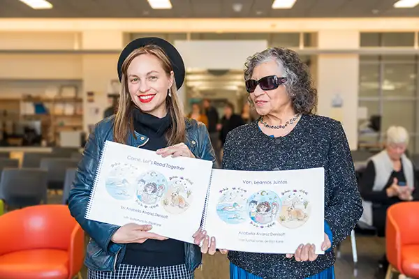And older and younger woman stand next to one another holding Spanish and English versions of the same children’s book