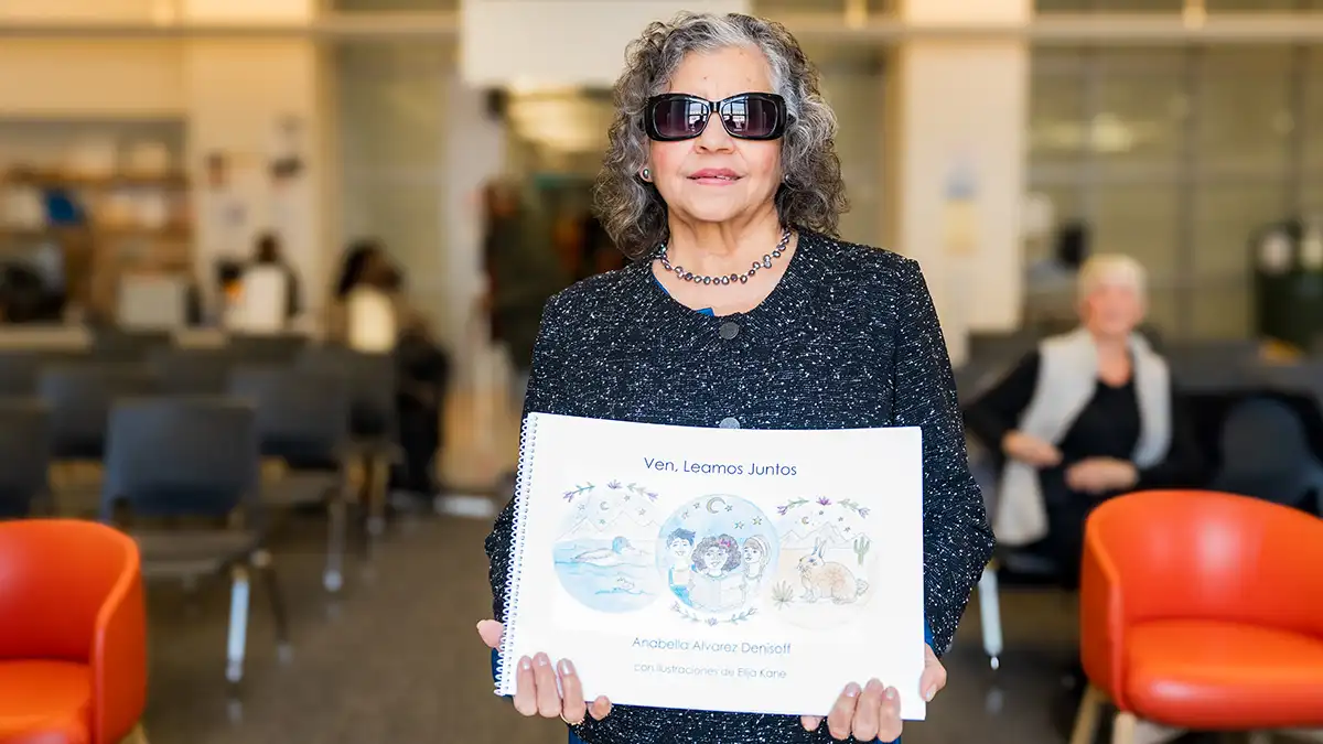 : An older woman in dark glasses holds a copy of an illustrated book in Spanish and Braille