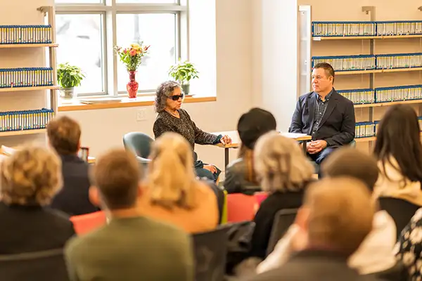 : A blind woman sits with a man in front of an audience in a library