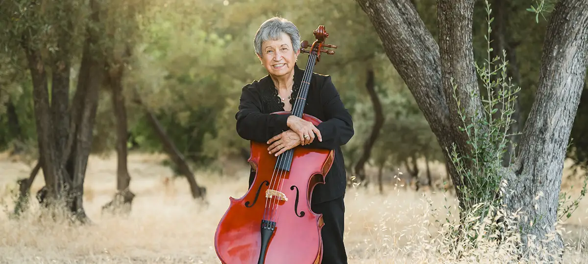 n older woman holding a cello stands outdoors among large trees.