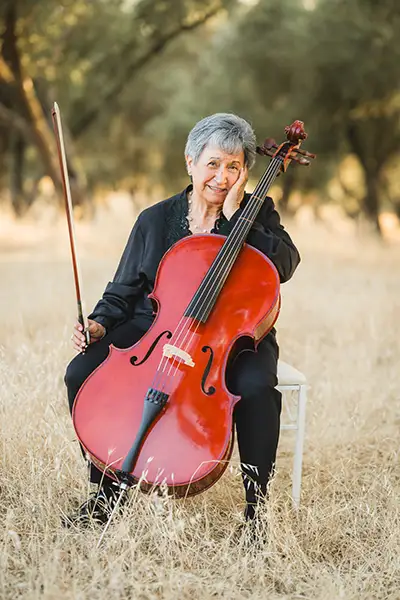 A smiling woman sits outdoors with a cello with a hand on her face.