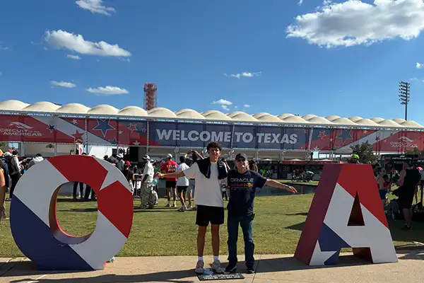 A teen boy and an older man stand in front of a race track stadium.