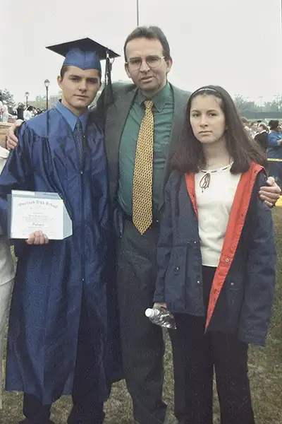 A father stands with his teen daughter and son who is wearing a graduation cap.