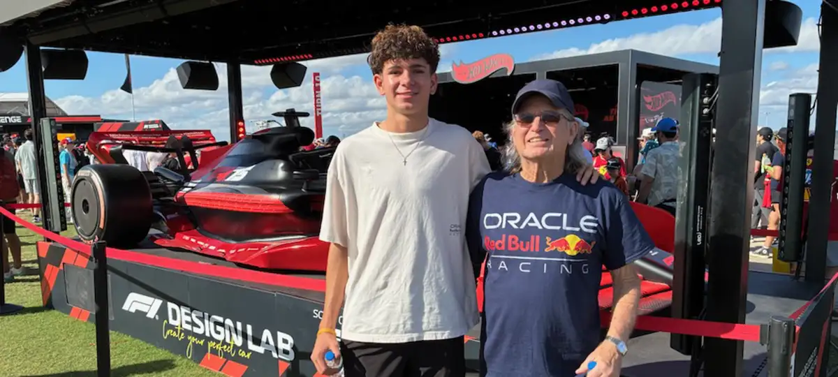 An older man stands arm in arm with a teen boy in front of a race car.