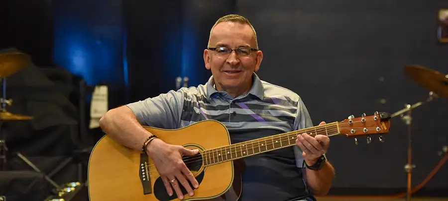 An older man with glasses holds an acoustic guitar.