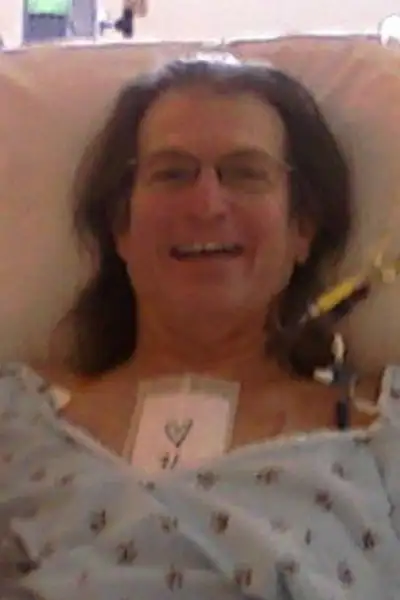 A man smiles in a hospital gown with a bandage on his chest.