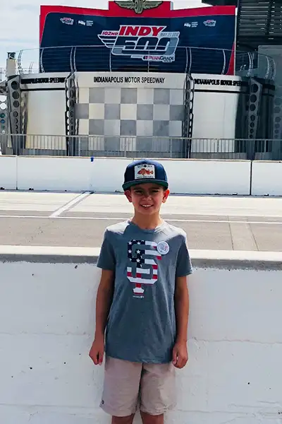 A young boy smiles in front of the Indianapolis 500 racetrack.