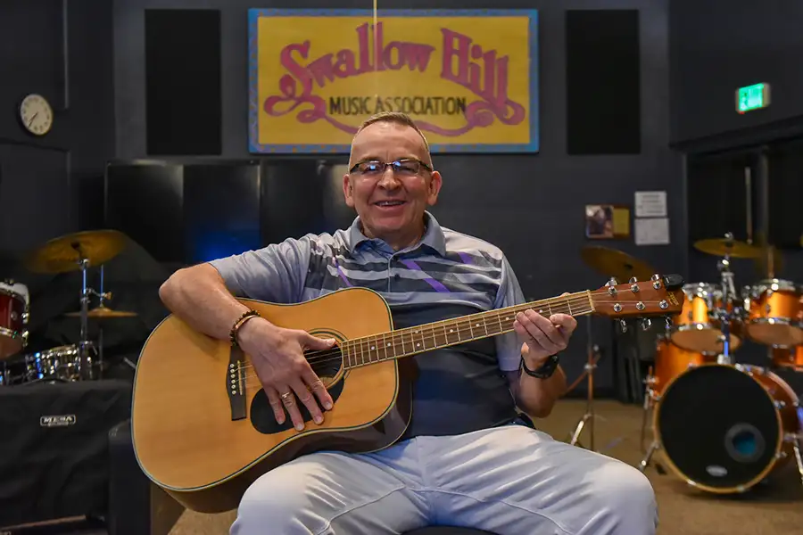 An older man holds a guitar in front of percussion instruments at a music center.
