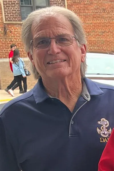 An older man in a collared shirt smiles in front of a brick building.