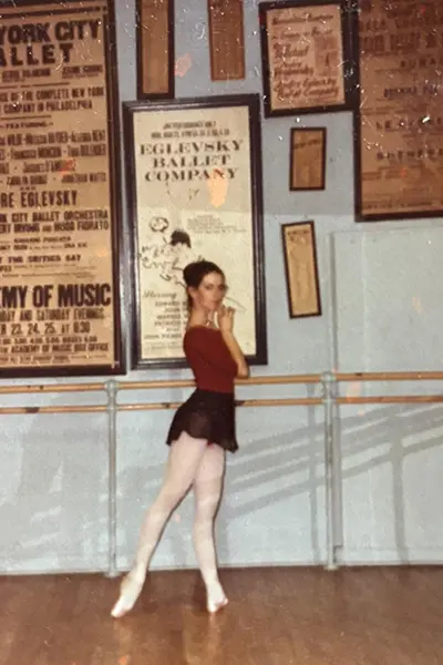 A dancer in pointe shoes stands along a barre.