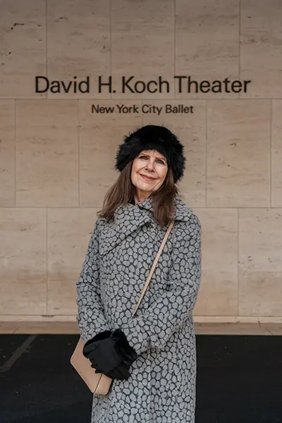 A woman in a coat and hat stands in front a New York City Ballet sign.